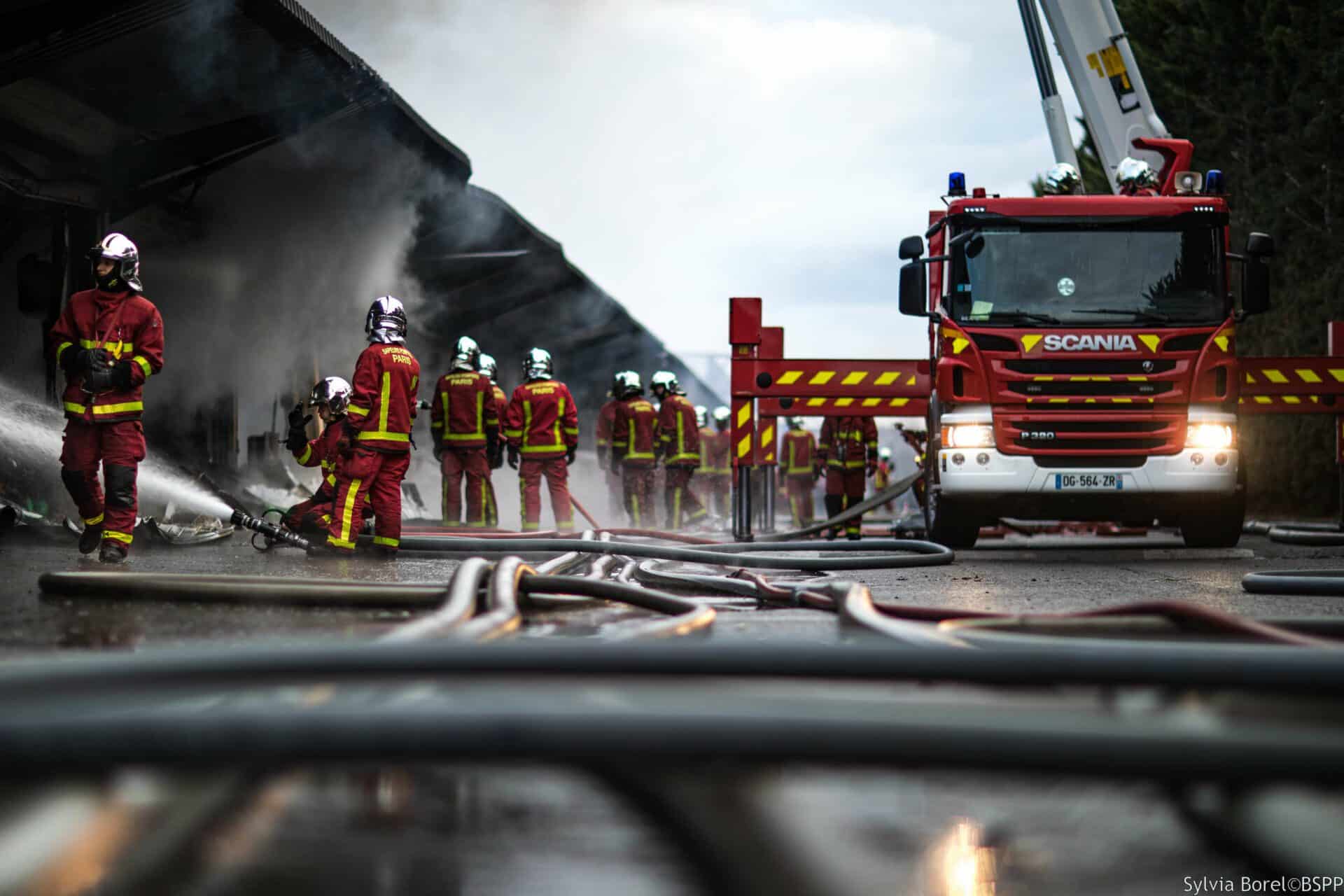 vu à ras du sol au niveau de tuyaux d'incendie, à quelques vingtaine de mètre, une dizaine de pompiers en tenue de feu, à droite un bras élévateur aérien
